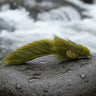 An olive-colored Woolhead Sculpin fishing fly resting on a bed of stones.
