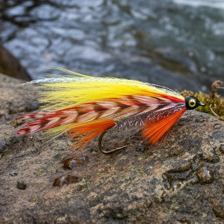 A close-up of a fly fishing lure on a textured off-white background. The lure features a silver glitter body, a black head with a yellow eye, a hook, and feathers in bright orange, red, and yellow.