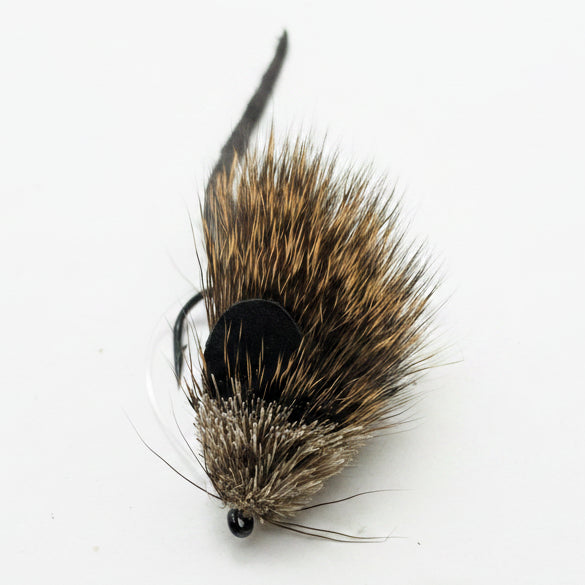 A close-up of a brown and black artificial fly fishing lure with a fuzzy body, black bead head, and dark tail, resting on a plain off-white surface.