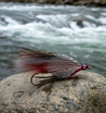 The image shows the Red-Grey Ghost Hairwing Variant streamer fly, with details about its construction including the use of quality 9X streamer hooks, red snow runner and dun cashmere goat wing, mirror flash and holographic flashabou, flat braided body in red, and stick-on eyes with UV over.