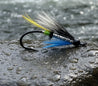 Fishing fly with black hook, blue body, and multicolored feathers on a light gray background